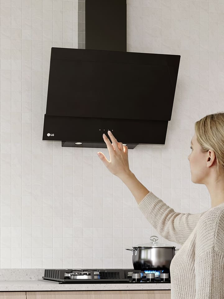 A woman adjusting the touch controls of a black glass hood above a cooktop in a modern kitchen with a white tile wall.
