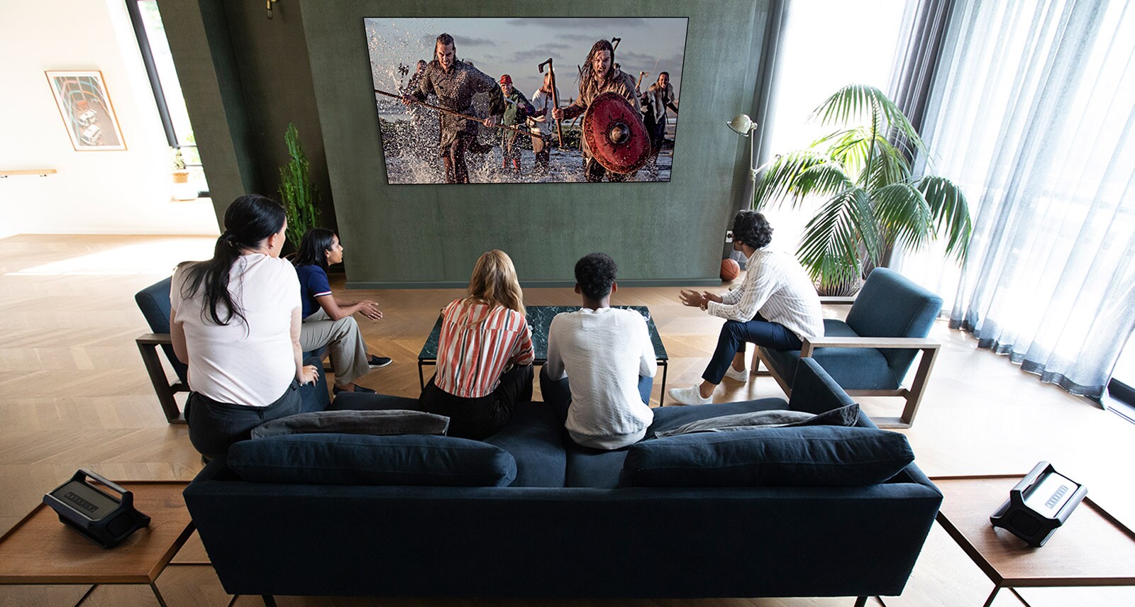 A group of friends watching a war movie on TV in the living room with 2 same Bluetooth speakers at rear speaker position