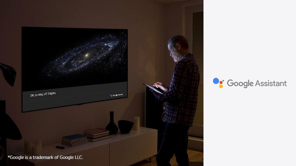 A man is standing in front of TV and ask google assistant to turn off the light
