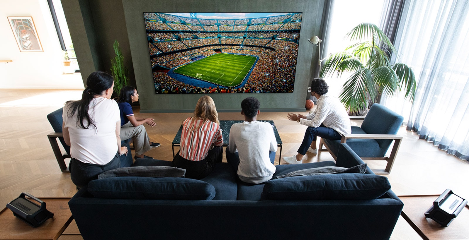 A group of friends watching a sports game on TV in the living room with 2 same Bluetooth speakers at rear speaker position