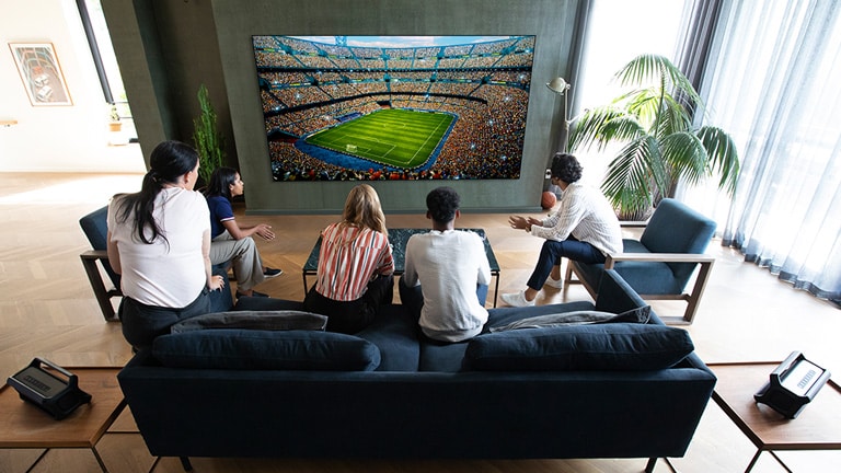 A group of friends watching a sports game on TV in the living room with 2 same Bluetooth speakers at rear speaker position