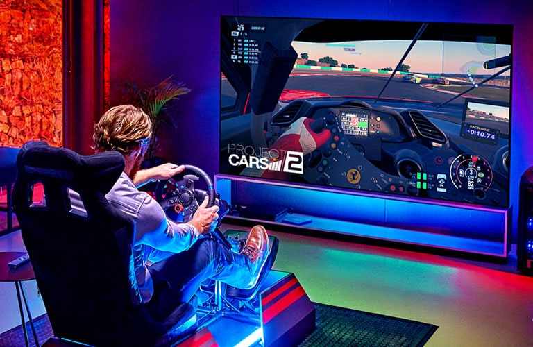 Man in a room sitting on a racing seat in front of a large TV playing a racing game 