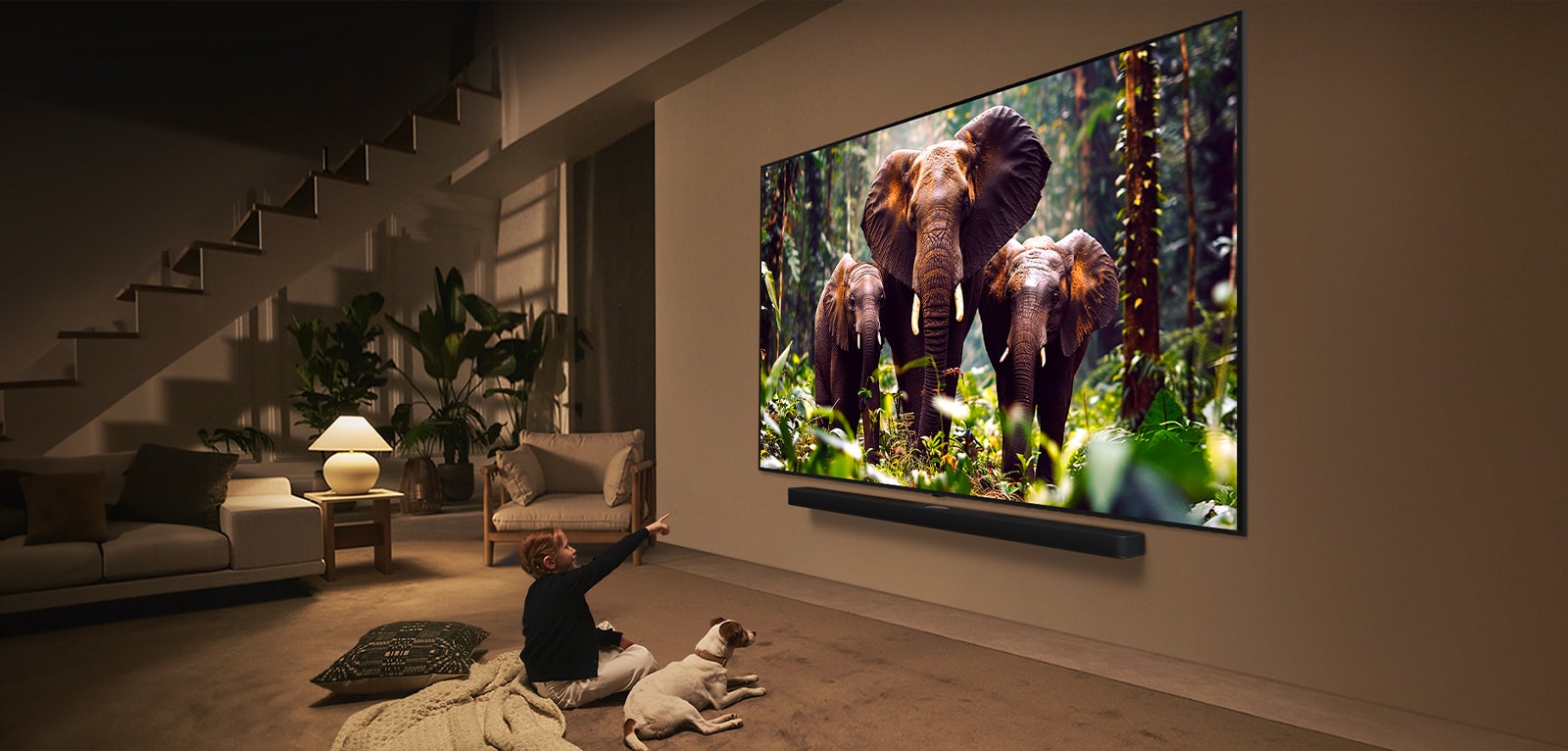 A girl and a dog are seated in front of a LG QNED TV mounted on a wall depicting three elephants walking outwards above a LG Soundbar. 