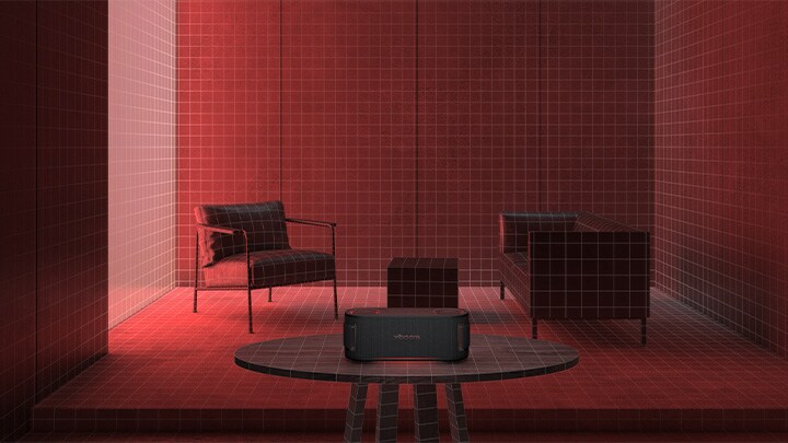 In a room filled with a red lighting a black sofa, a black ottoman  and a black armchair are placed behiind. In the fornt, xboom Bounce is placed on the top of black round table.
