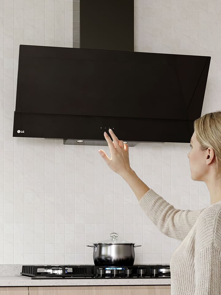 A woman adjusting the touch controls of a black glass hood above a cooktop in a modern kitchen with a white tile wall.