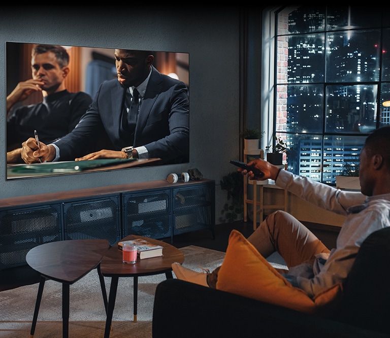 A man is sitting on the sofa watching TV. The man has a remote control in his hand, and TV shows a scene from the movie.