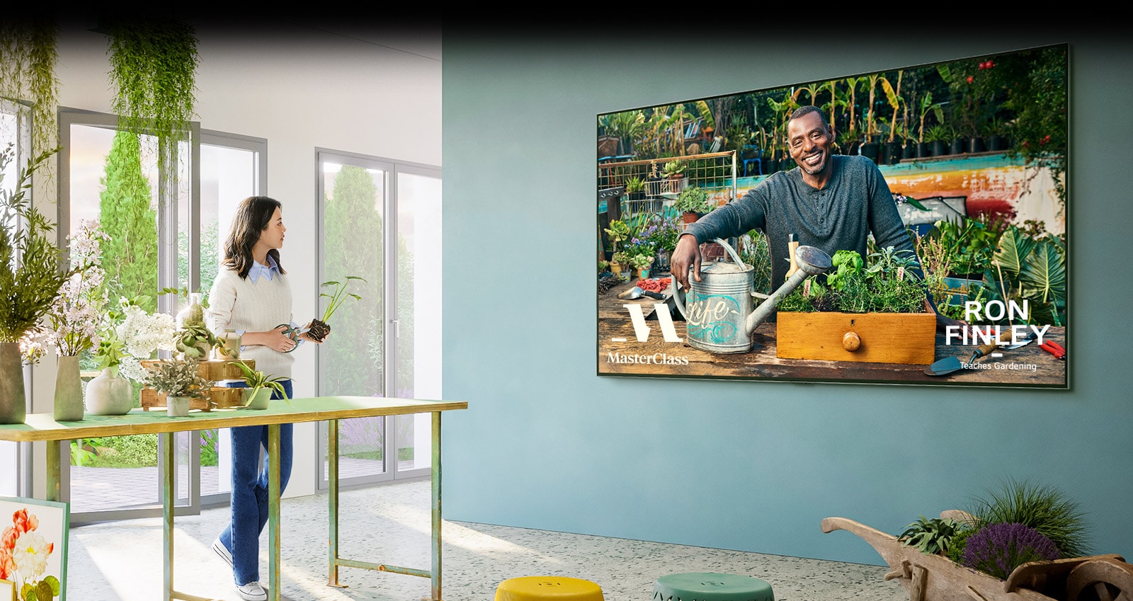 A large TV is on the wall, and the TV shows the gardening class screen of 'Master Class'. A woman is standing at a table next to a TV and taking a gardening class with flower pots and flower scissors.