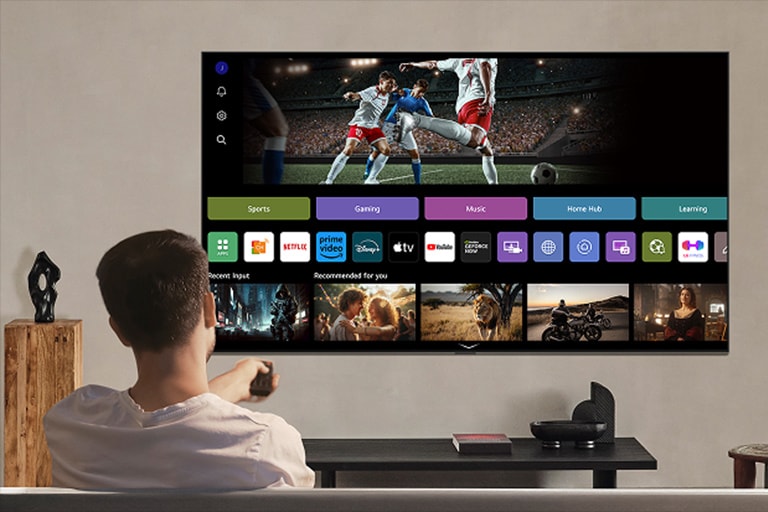 Person on the couch holding out a remote control. LG AI TV on the wall with LG webOS on the screen.