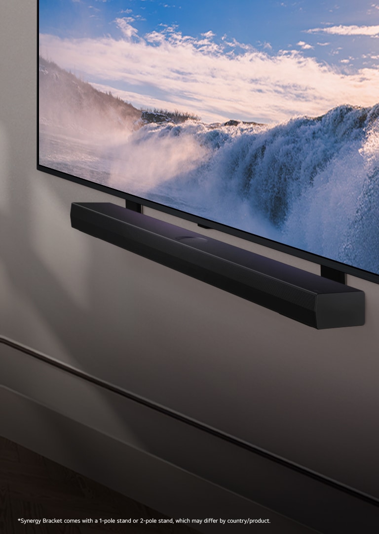 LG TV and Soundbar are placed within an angled perspective mounted on a wall. On the TV, a close up of a vast waterfall is displayed, and soft sunlight cascade over the wall, TV, and soundbar. A disclaimer reads: "Synergy Bracket comes with a 1-pole stand or 2-pole stand, which may differ by country/product."