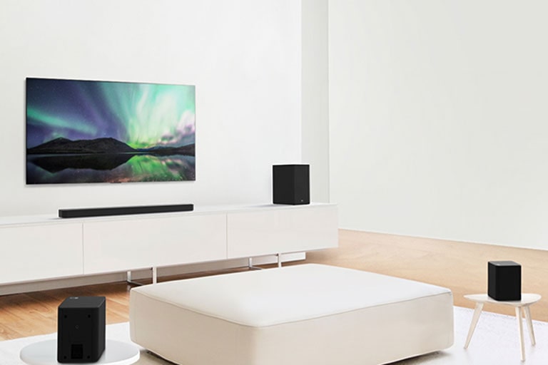 TV and sound bar in white living room with white sofa in the center. Speakers sit on both ends of the sofa.