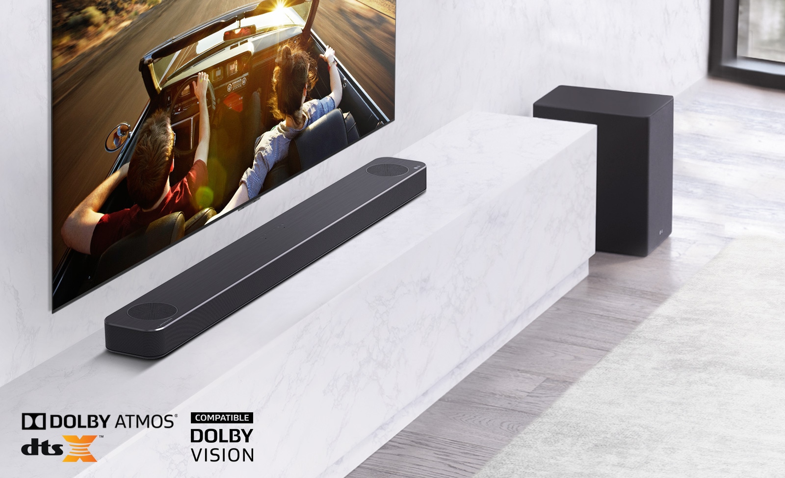 TV is on the wall, LG Soundbar is below on a white marble shelf with a sub-woofer to the right. TV shows a couple in a car. 