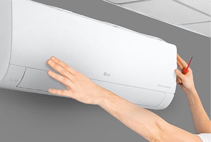 The side view of the air conditioner can be seen on the wall. Two hands are reaching up, one holding a tool, showing the ease of installation.