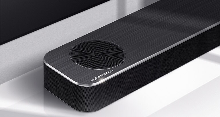 Primer plano del lado izquierdo de la barra de sonido LG con el logotipo de Meridian en la esquina inferior izquierda del producto. También puede verse la parte inferior izquierda de la TV. 