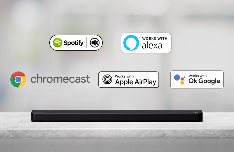 Hay una barra de sonido ubicada en un estante gris y hay logos de plataformas de IA, en orden de Spotify, Alexa, Chromecast, Apple Airplay y OK Google de izquierda a derecha.