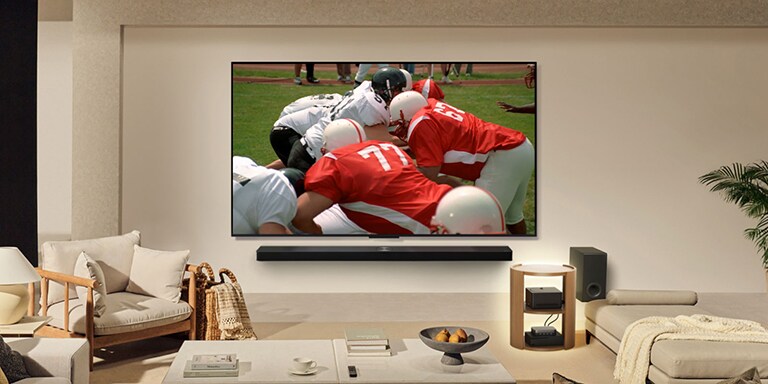 Una sala de estar con una LG True Wireless TV en la pared. En la pantalla se ve un partido de fútbol americano. La instalación de la TV también incluye una LG Soundbar montada en la pared. Y, casi imperceptibles, el resto de los dispositivos periféricos están conectados a una Caja de conexión cero que se encuentra sobre una mesa de apoyo. La sala de estar está prolija, ordenada y no hay desorden de cables.