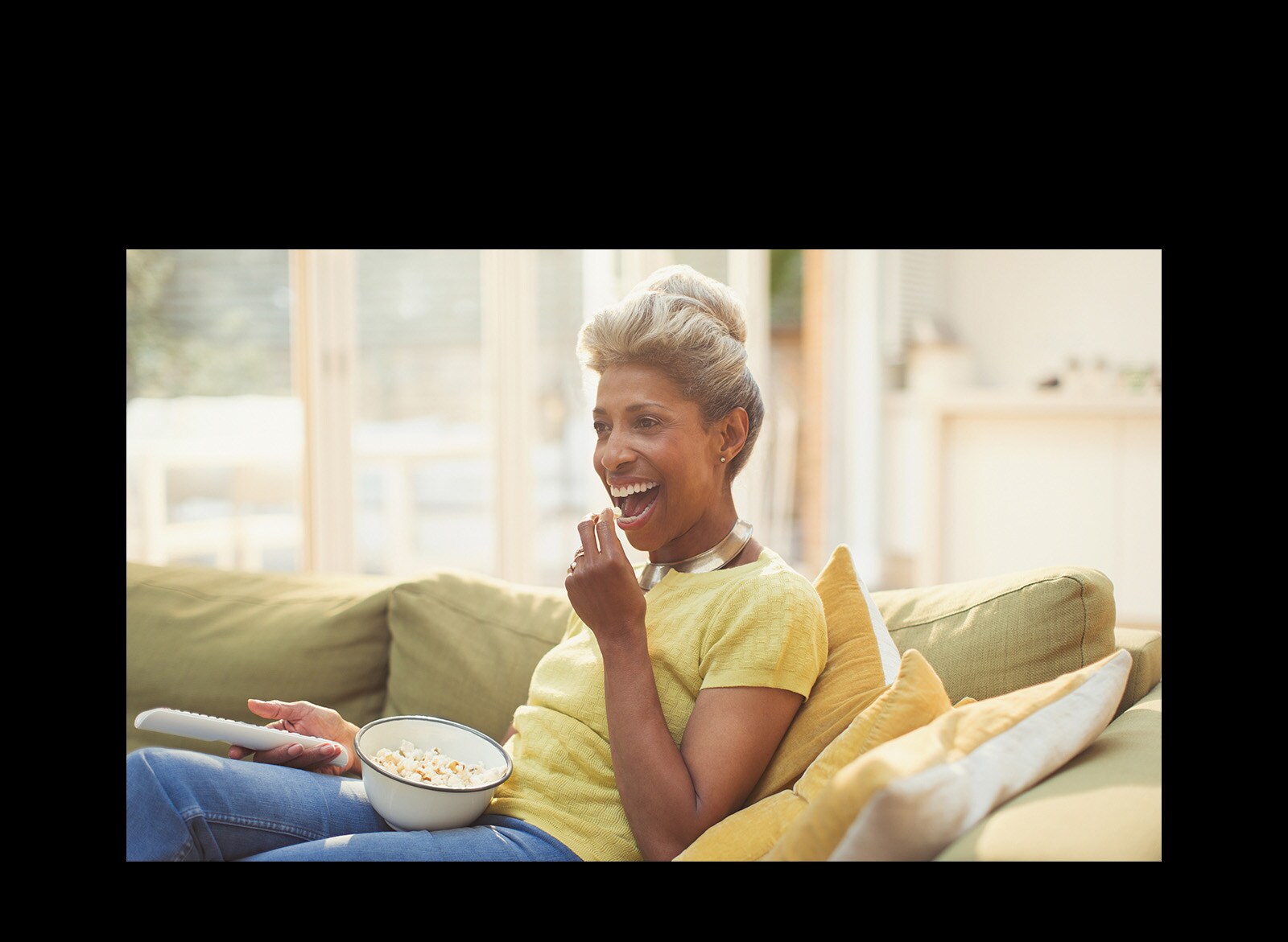 Una mujer está viendo la televisión, y sostiene un control remoto. También está comiendo palomitas de maíz.