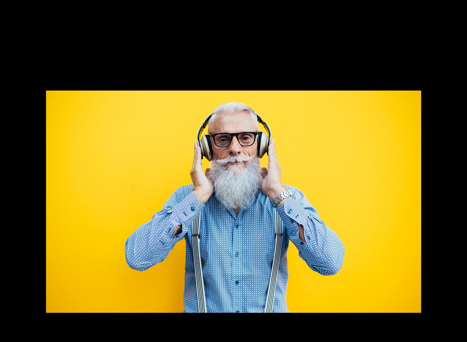 Un abuelo está parado frente a una pared amarilla, con auriculares.
