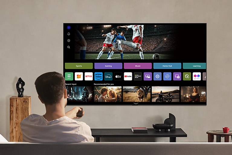 Una persona en el sofá con un control remoto en la mano. LG AI TV en la pared con LG webOS en la pantalla.