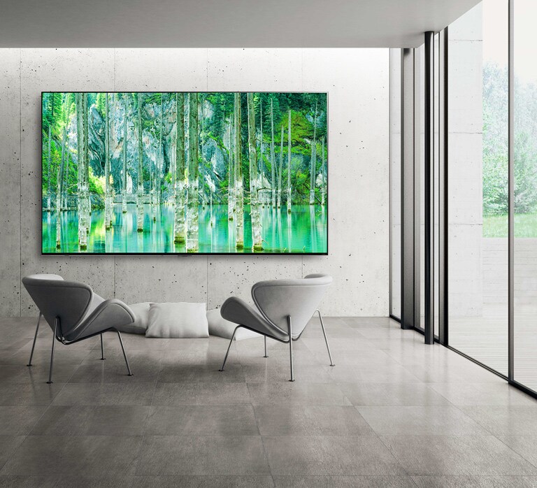 Una imagen de un televisor LG QNED MiniLED de pantalla grande montado en una pared gris de cemento sin detalles decorativos. Hay dos sillas dispuestas frente al televisor y en la pantalla se ve una escena natural de color verde.