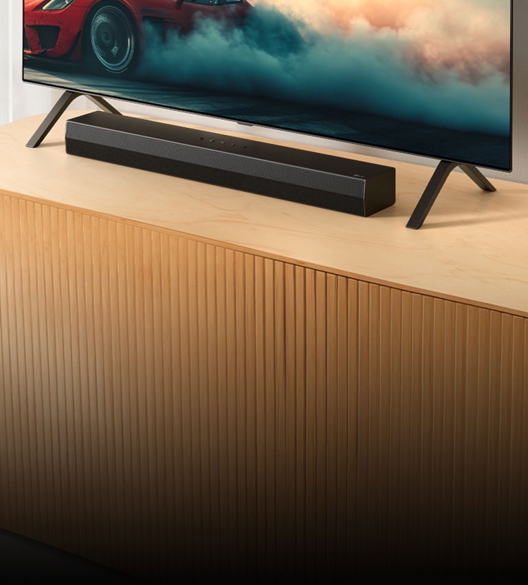 La barra de sonido LG S20A se encuentra sobre una consola de madera para TV, debajo de un televisor y entre sus soportes. En el televisor se muestra un automóvil rojo con humo.