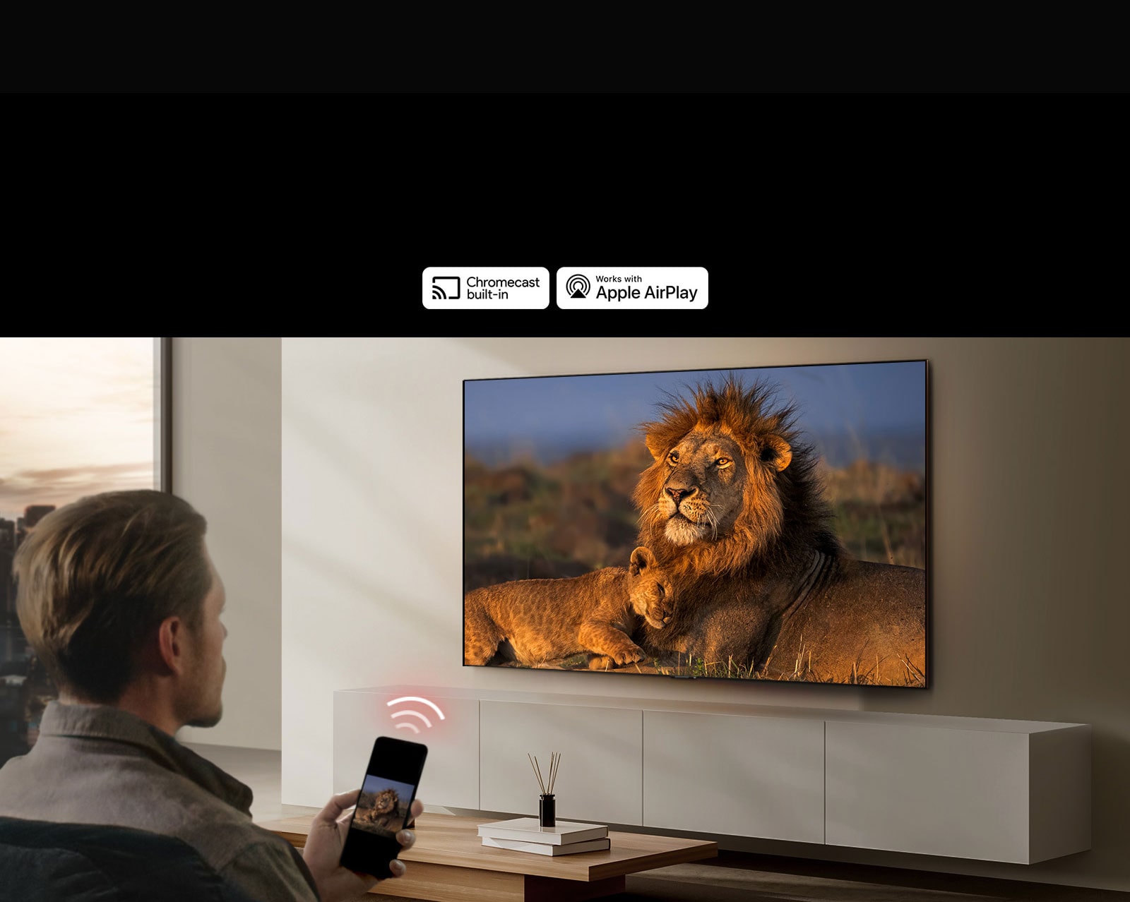 An LG TV mounted on a wall in a living room, displaying a lion and lion cub. A man sits in the foreground with a smartphone in his hand displaying the same image of lions. A graphic of three neon red curved bars is displayed just above the smartphone pointing toward the TV.