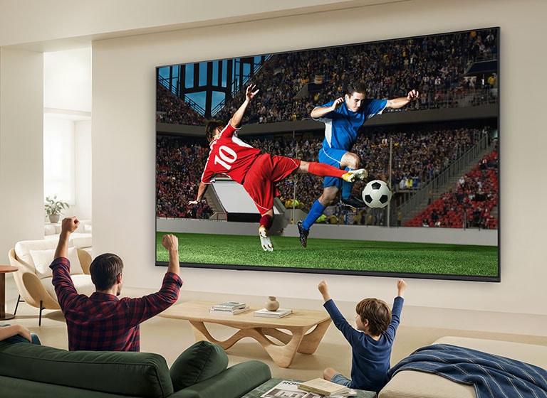 LG QNED AI Mini LED QNED70 Ultra Big TV on elutoas seinale paigaldatud ning kuvab intensiivset jalgpallihetke erksate värvide ja viimistletud pildikvaliteediga laial ekraanil, samal ajal kui diivanitel istuv perekond koos juubeldab.