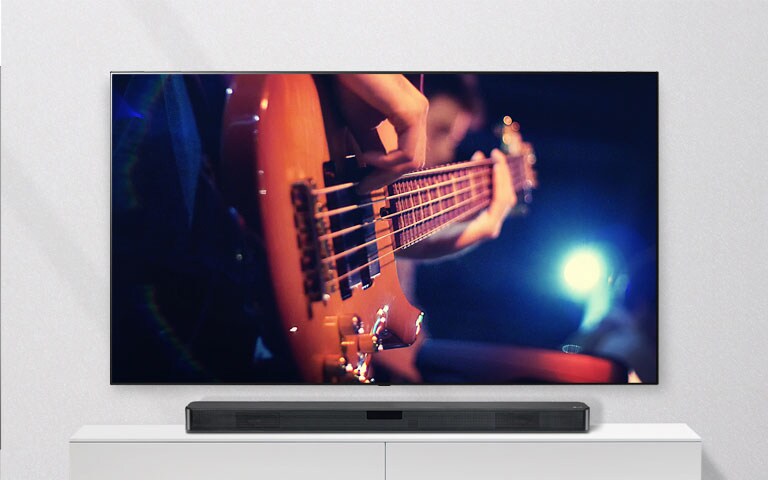 Teler on seina külge kinnitatud ja heliriba on valgel riiulil. Teler näitab kitarri mängivat meest.