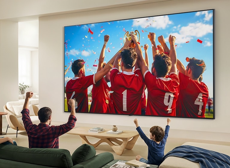 LG QNED evo AI QNED85 Mini LED Ultra Big TV on elutoas seinale paigaldatud ning kuvab spordivõidu tähistamist erksate värvide ja viimistletud pildikvaliteediga laial ekraanil, samal ajal kui diivanitel istuv perekond koos juubeldab.