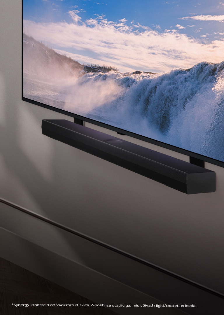 LG teler ja Soundbar ribakõlar on seinale kinnitatud nurga all. Teleris on kuvatud lähivõte suurest kosest ning pehme päikesevalgus voogab üle seina, teleri ja ribakõlari. Kohustustest loobumise avalduses on kirjas: **Synergy kronstein on varustatud 1-või 2-postilise statiiviga, mis võivad riigiti/tooteti erineda.