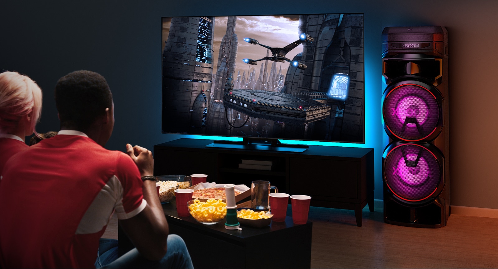 Inimesed vaatavad telerit, mis on paigaldatud seinale ja millest paremal on kõlar LG XBOOM RNC7. Teleris mängib film.