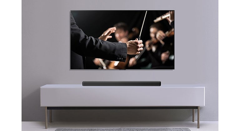 A TV is shown on a gray wall and LG Soundbar below it on a gray shelf. TV shows a conductor conducting an orchestra. 