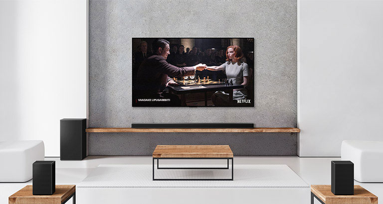 2 tagumise, bassi- ja soundbari ning teleri komplekt on valges elutoas. Naine ja mees mängivad teleriekraanil malet.