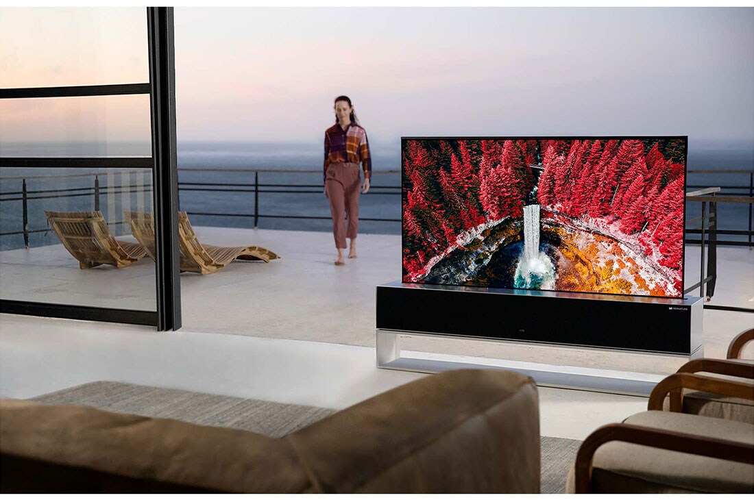 LG 65-tolline OLED 4K teler, Elutuba koos kokkurullitava teleriga, mis näitab vahtrate vahel asuvat koske, samal ajal kui terrassilt siseneb tuppa naine, OLED65RX9LA, thumbnail 11