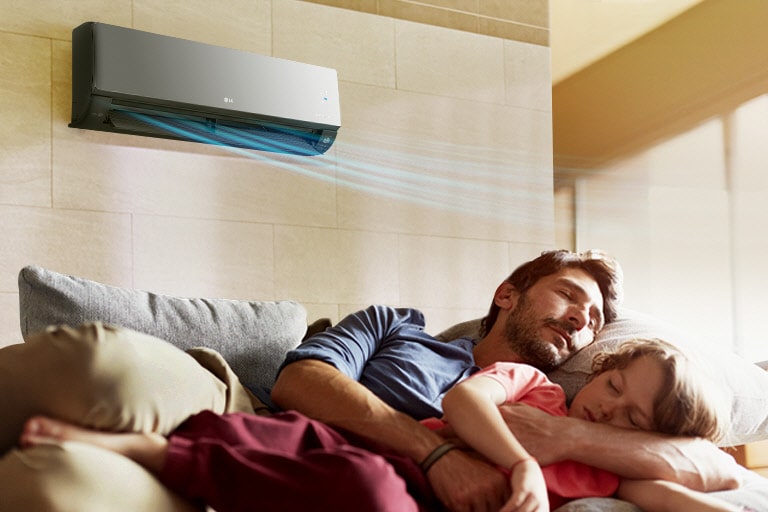 A father and daughter sleep on a couch beneath an air conditioner that is blowing air out over them.