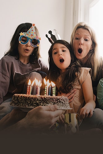 Image of two adult women and a young girl wearing a birthday hat on their head and blowing out the candles on the cake.