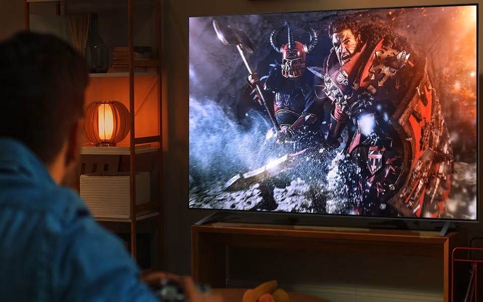 Una persona juega a un videojuego en un televisor LG. En la pantalla, dos guerreros con armadura luchan en una escena intensa, mientras una lámpara ilumina la habitación con una luz cálida.