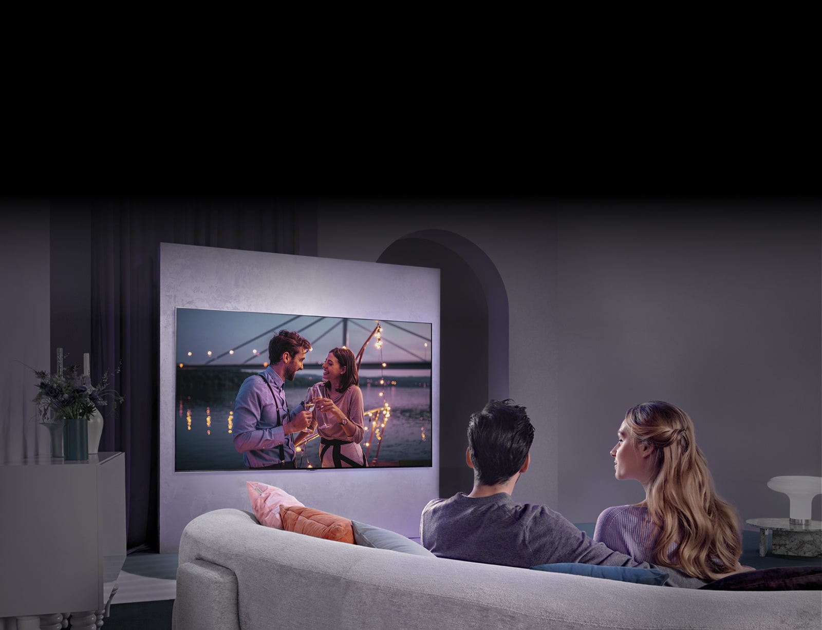 Henkilö istuu sohvalla ja katselee elokuvaa, joka näkyy suurella televisiolla seinällä.