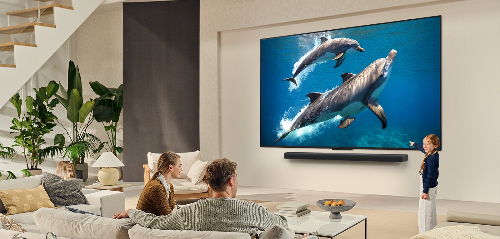 Perhe istuu sohvalla katselemassa seinälle asennettua LG QNED TV -televisiota, jonka alapuolella on LG Soundbar. Pikkutyttö osoittaa ruutua, jossa näkyy kaksi delfiiniä. 
