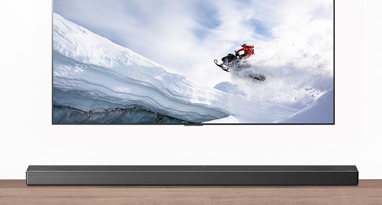 Televisio ja soundbar-kaiutin edestäpäin. Televisiossa mies ajaa lumikelkalla lumisilla vuorilla. HDMI 2.1 -logo television alapuolella. 