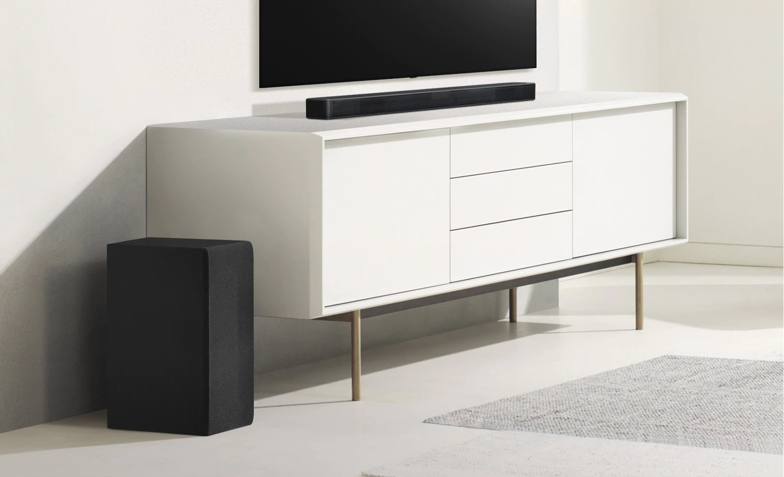 Soundbar-kaiutin on valkoisessa hyllyssä, ja vasemmalla puolella on subwoofer. Subwoofer näyttää suuremmalta, koska se on edempänä.