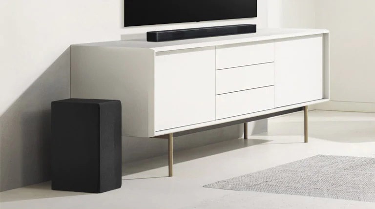 Soundbar-kaiutin on valkoisessa hyllyssä, ja vasemmalla puolella on subwoofer. Subwoofer näyttää suuremmalta, koska se on edempänä.