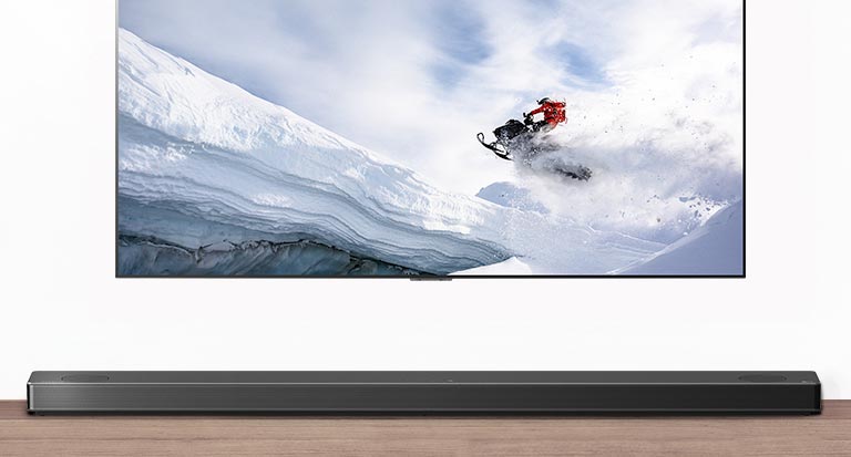 Televisio ja soundbar-kaiutin edestäpäin. Televisiossa mies ajaa lumikelkalla lumisilla vuorilla. HDMI 2.1 -logo television alapuolella. 