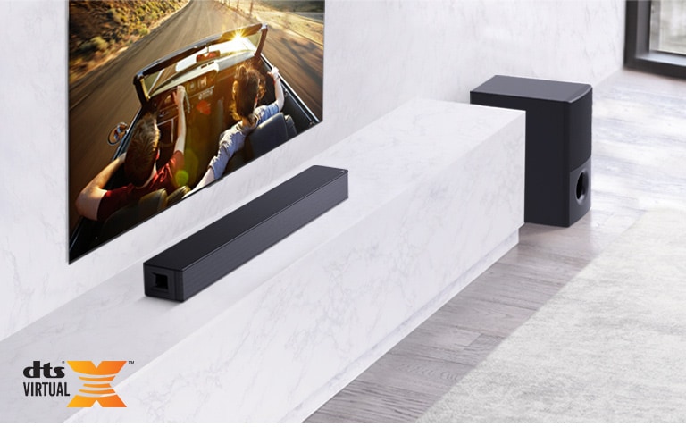 Televisio on seinällä. LG-soundbar-kaiutin on sen alapuolella valkoisen marmoritason päällä, ja kaiuttimen oikealla puolella on subwoofer. Televisiossa näkyy autossa oleva pariskunta.