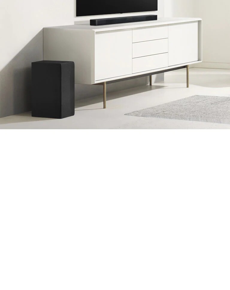 Soundbar-kaiutin on valkoisessa hyllyssä, ja vasemmalla puolella on subwoofer. Subwoofer näyttää suuremmalta, koska se on edempänä.