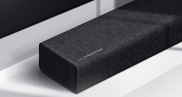Lähikuva LG Soundbar -kaiuttimen vasemmasta reunasta, jonka vasemmassa alakulmassa näkyy Meridian-logo. Television vasen alareuna on myös näkyvissä.