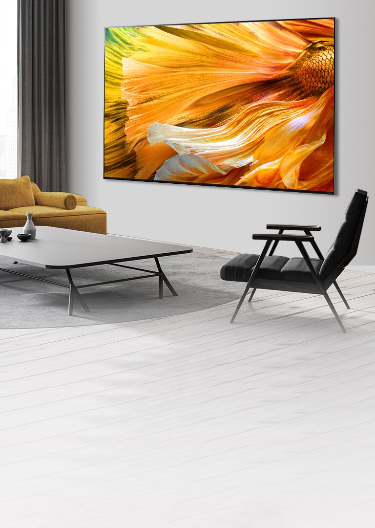 Kuva suuren kuvaruudun LG QNED MiniLED -televisiosta, joka on asennettu harmaalle seinälle.  Television eteen on aseteltu sohva, sohvapöytä ja tuoleja.