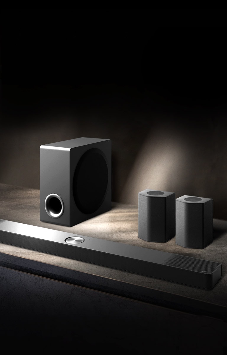 LG Soundbar, takakaiuttimet ja subwoofer on asetettu kulmasta näkyvään ruskeaan puuhyllyyn mustassa huoneessa, jonka ainoa valaistus kohdistuu äänijärjestelmään.