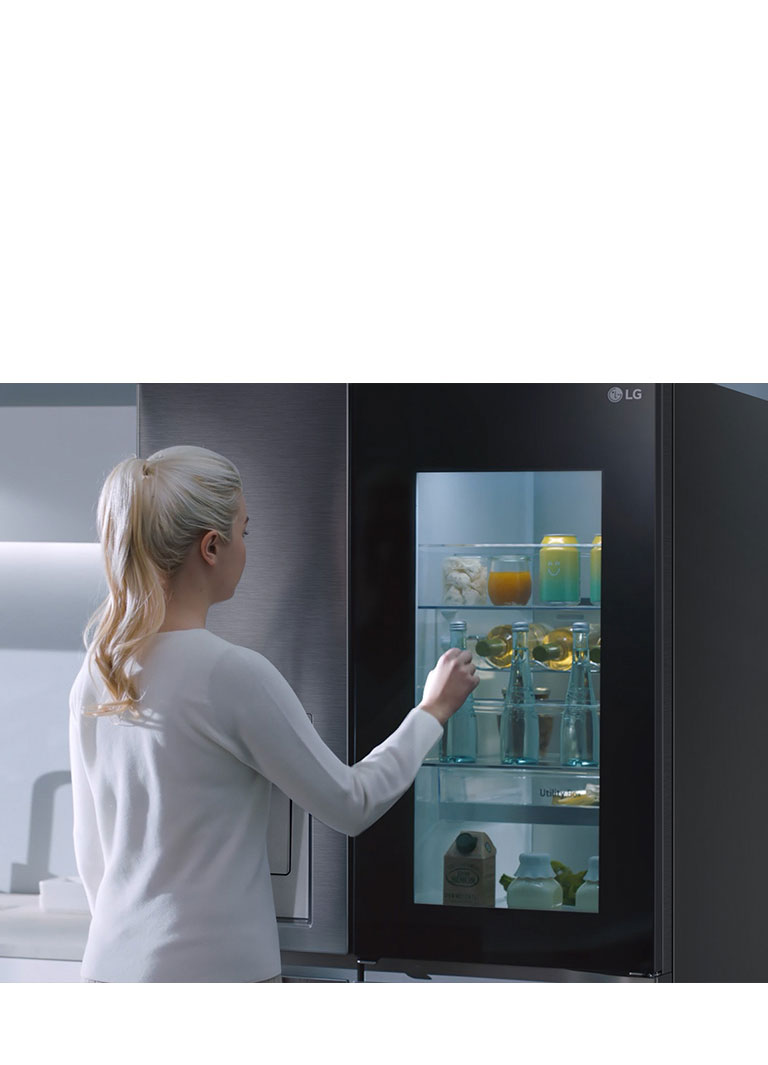 A video shows a woman approach her InstaView refrigerator and knock twice. The interior lights up and she can see the contents of her fridge without opening the door. The view zooms in to focus on the drinks in the door and then zooms out to see the woman from behind as she opens the door and grabs a drink.