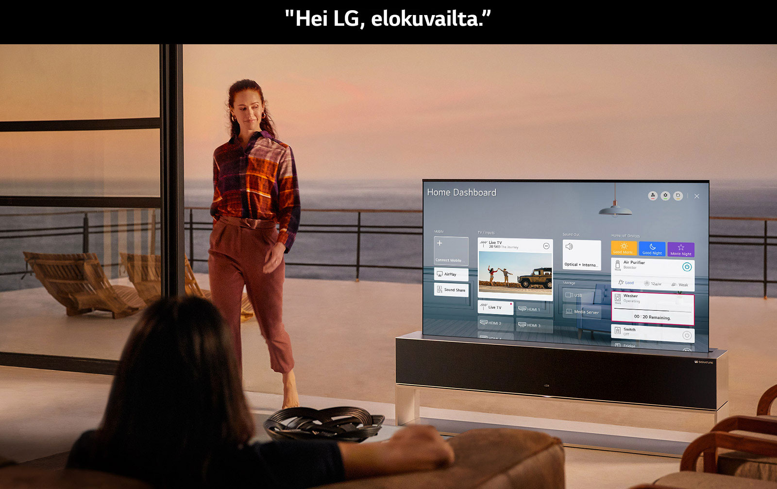 A woman viewing the Home Dashboard on TV in the living room with an ocean-front terrace, while another woman walks inside