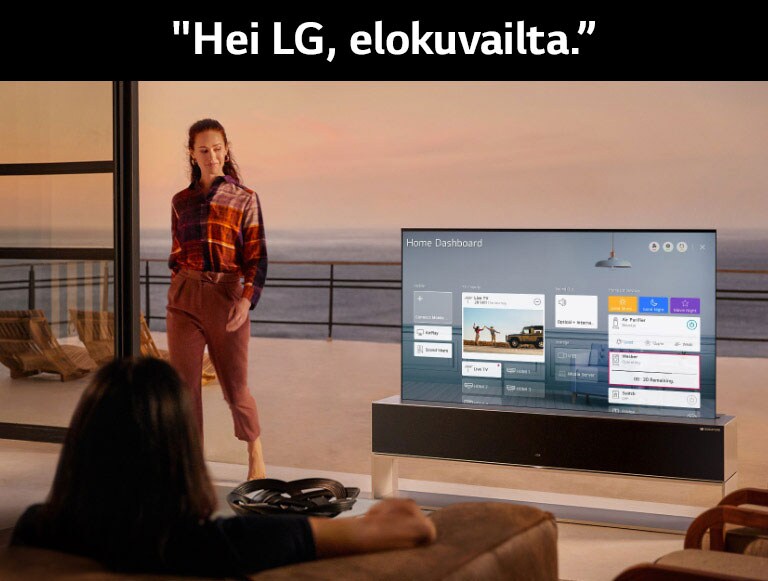 A woman viewing the Home Dashboard on TV in the living room with an ocean-front terrace, while another woman walks inside